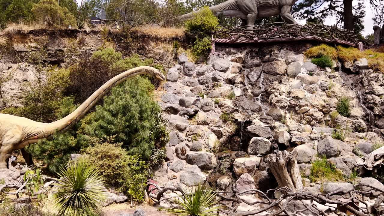 Close-up of a long-necked dinosaur statue looking at the camera with a small waterfall in the background and another dinosaur behind it