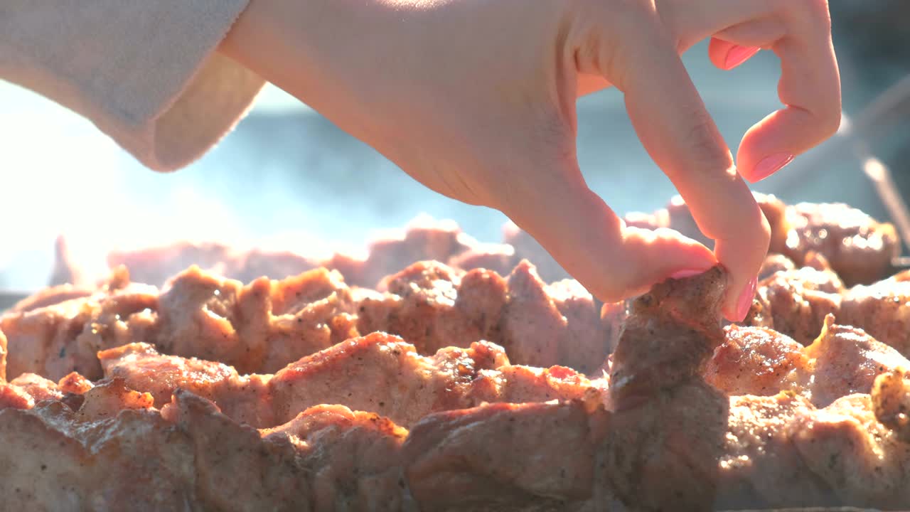 Woman tears a piece of meat pork barbecue roasted on skewers on top of charcoal grill. Pieces of meat and woman's hand close-up.
