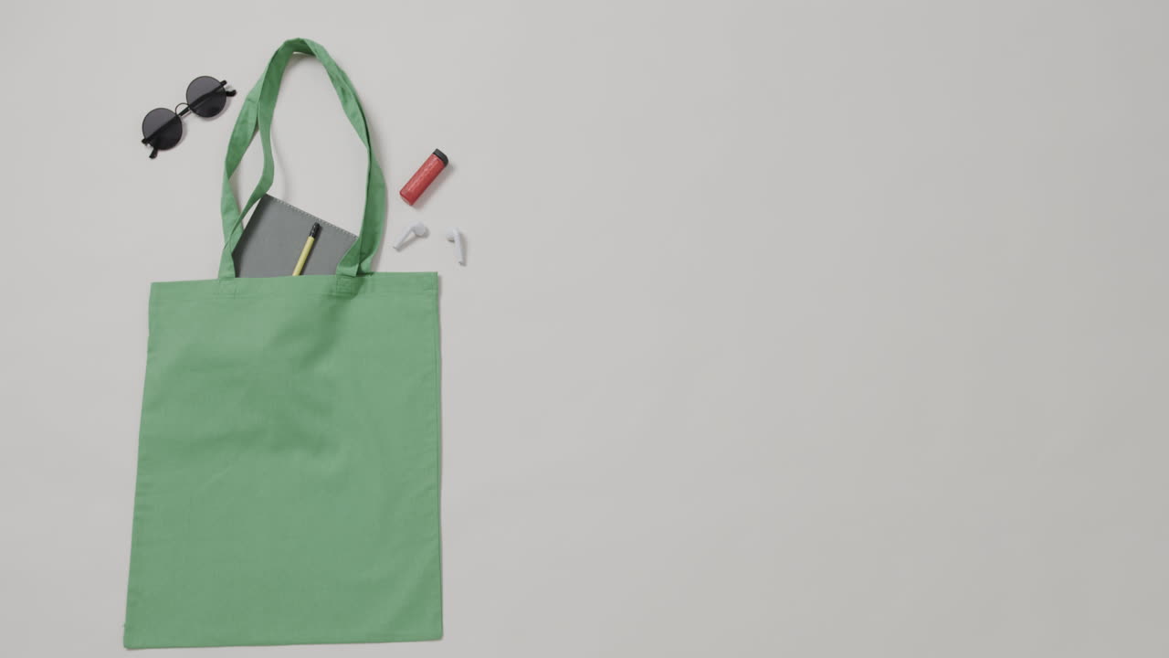 bolsa verde con gafas de sol, cuaderno, lápiz labial sobre fondo blanco, espacio para copiar, cámara lenta