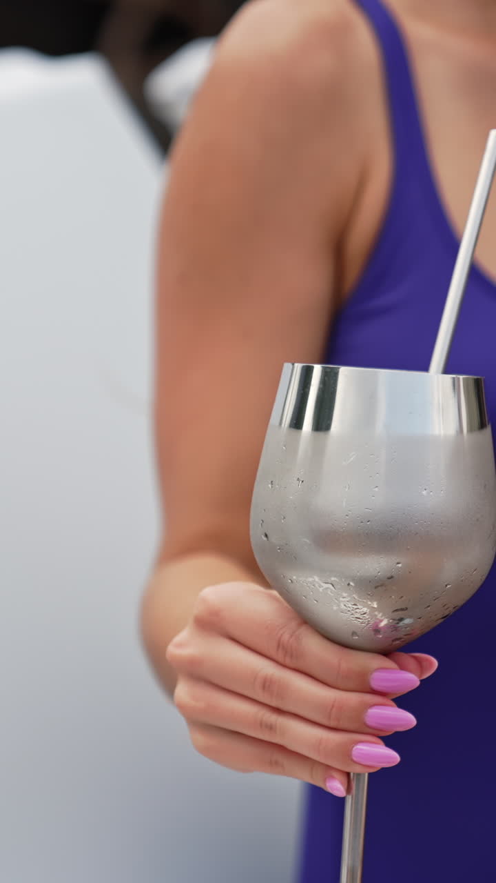 Metal glass with a straw taken by the female hand. Young dark-haired lady drinking cocktail sitting on a deck chair. Vertical video