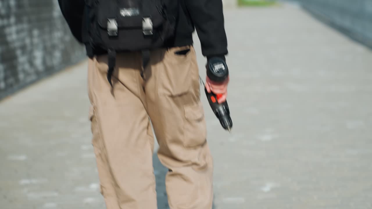 White man walking with drill along city bridge, tan cargo pants, black backpack, wristwatch on left hand, steady stride, tense atmosphere suggesting covert technician or suspect, daytime light