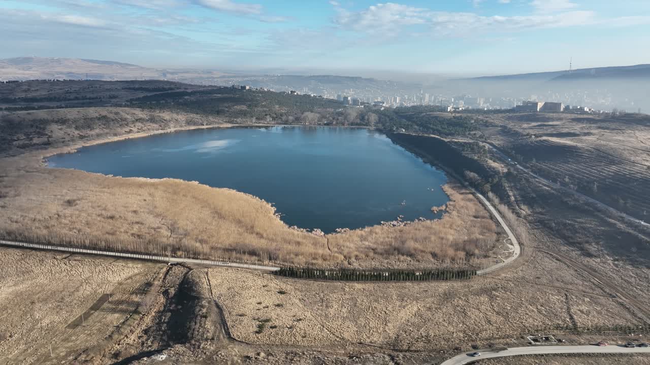 A secluded lake resting within a plateau, reflecting the sky. The reeds form a contrast between the golden land and the deep blue water, with distant city structures faintly visible on the horizon