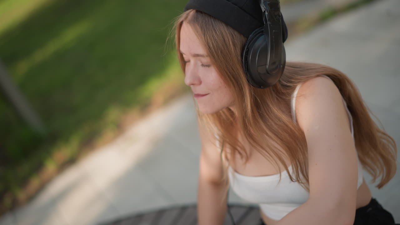 Giovane donna seduta su una panchina del parco che ascolta musica con cuffie over-ear e berretto. Latte macchiato freddo su un tavolino rotondo in legno accanto a lei. Luce del sole che filtra tra gli alberi, ombre leggere sul pavimento. Espressione rilassata