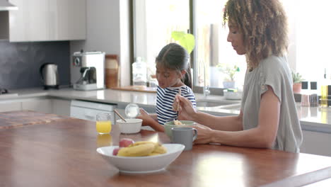 Madre-E-Hija-Birraciales-Comiendo-Cereales-Para-El-Desayuno-En-Una-Cocina-Soleada,-Espacio-Para-Copiar,-Cámara-Lenta