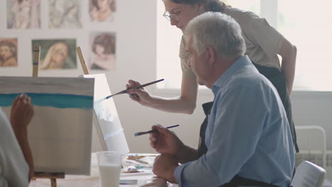 a female teacher shows a retired man how to draw a picture with paints and a brush at courses for the elderly. a senior man draws a picture to a group of pensioners