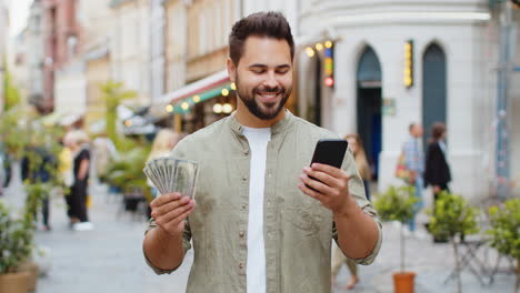 happy young man counting money dollar cash, use smartphone calculator app in urban city street