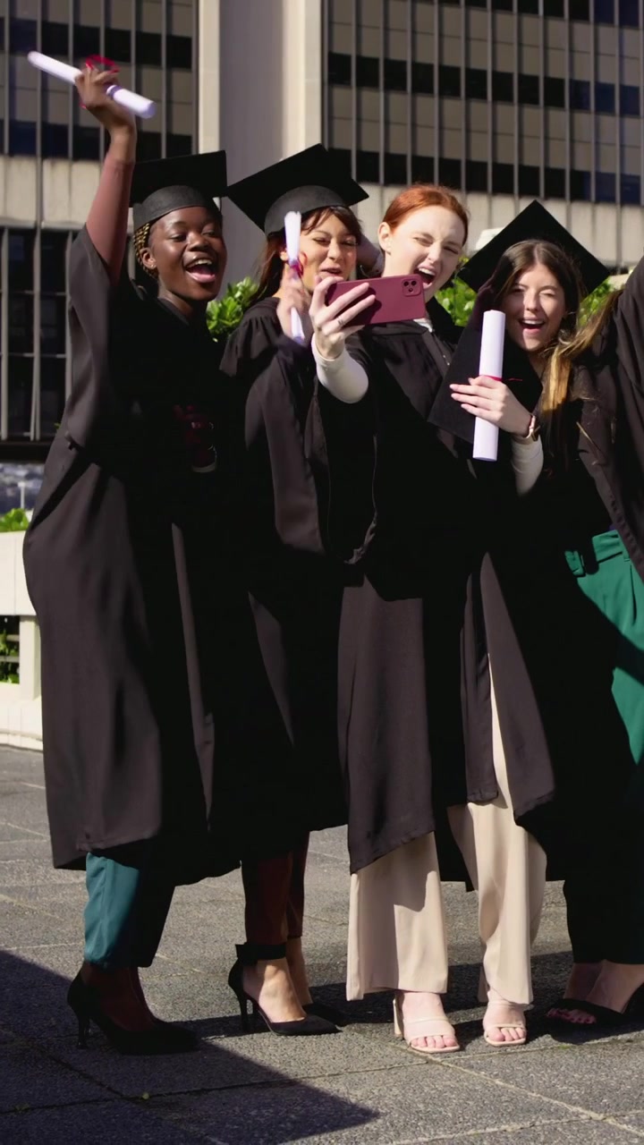 Video verticale: Tirando fuori il telefono, 4 laureati si scattano un selfie in piazza, festeggiando con diplomi e toghe.