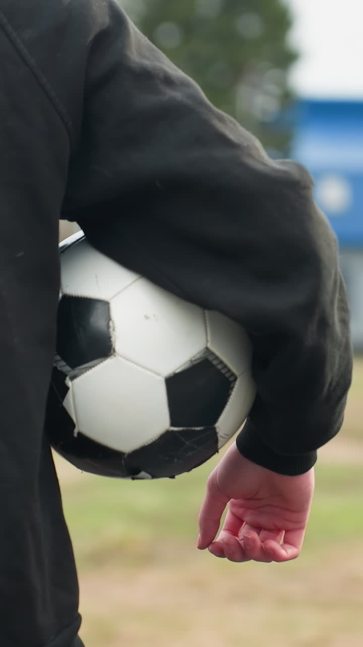 Back view of someone in a black outfit holding a classic black and white soccer ball under their arm, with a blurred grassy field and a blue building in the background