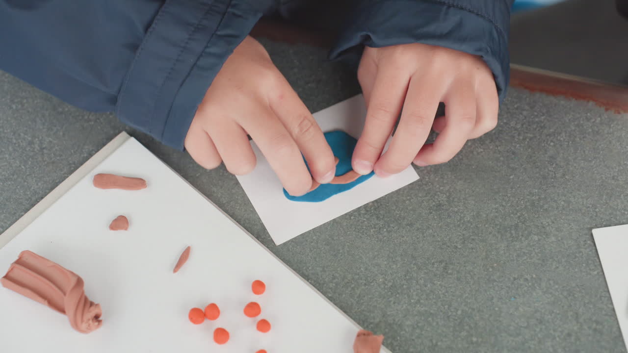 Student hands arranging clay pieces on card, assembling orange beads and brown strips into shape, workspace view with papers and tools, hands molding and organizing, playful classroom prep for back