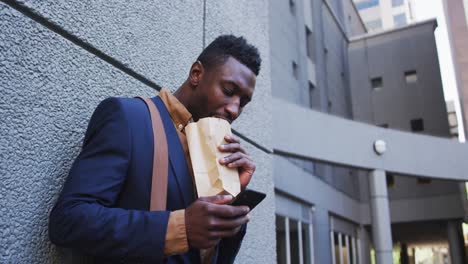 African-american-businessman-eating-sandwich-using-smartphone-in-city-street