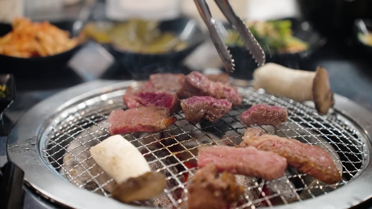 Chef uses metal tongs and kitchen shears to flip and slice pieces of sizzling Hanu beef on a hot kbbq grill with smoke rising in slow motion