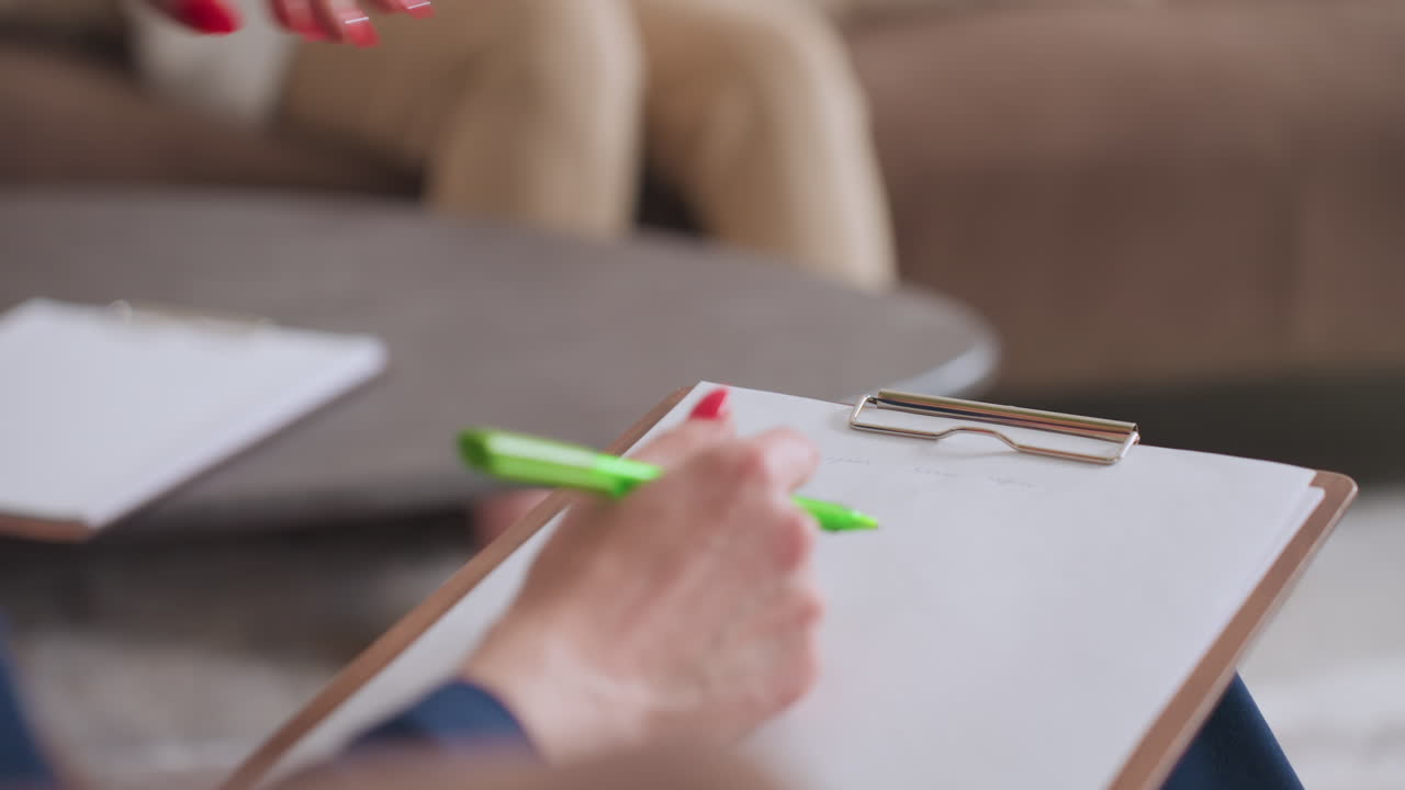 Close view of therapist hand holding green pen while writing on white clipboard during client session, seated comfortably near table, background includes blurred view of cozy setting