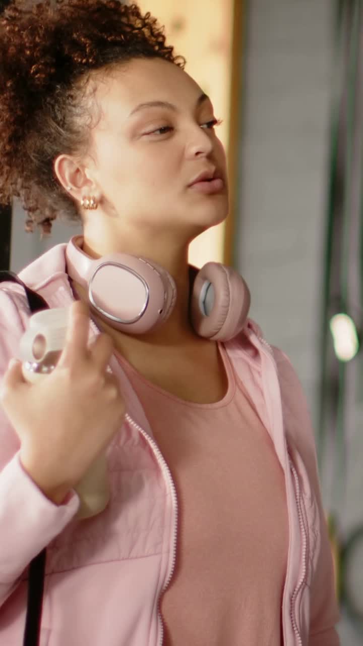 Video verticale: Sentendo la chiamata del partner, donna con cuffie rosa e giacca che parla, tenendo una bottiglia in studio.
