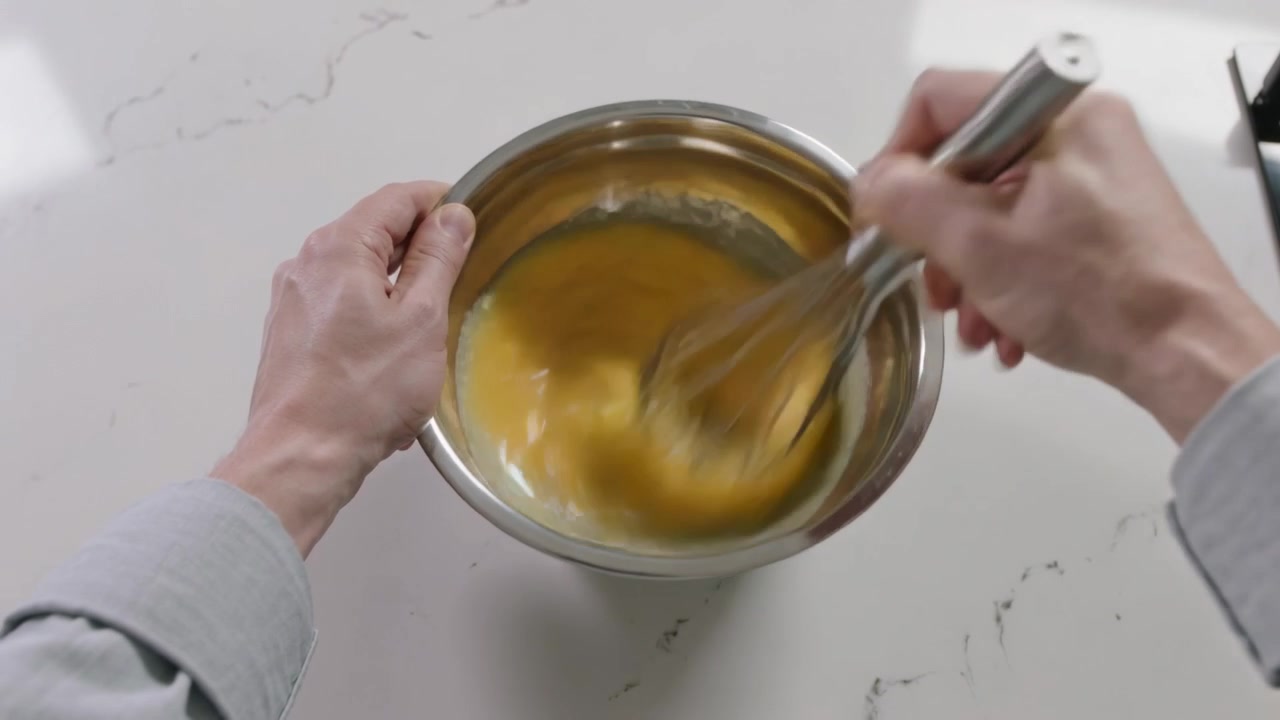 Sbattendo un uovo, mani con maniche lunghe che tengono una ciotola d'acciaio e una frusta sul bancone della cucina.