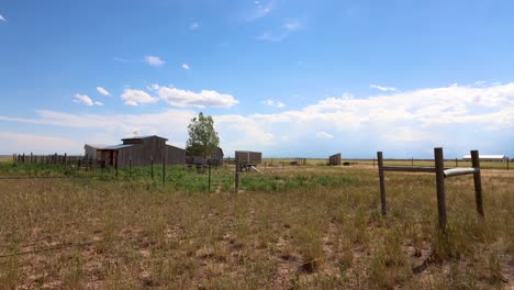 an old time western barn with a corral and weather vein stands century over a barren plain while puffy white clouds form and dissipate against a stark blue sky