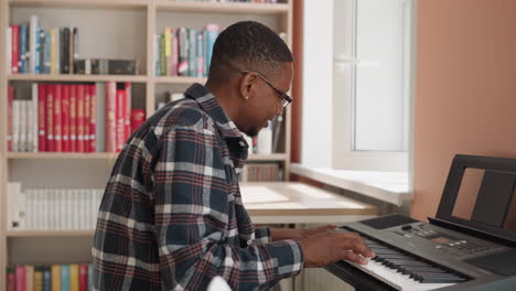 man presses keys of synthesizer by bookcase. smiling african american musician practices tune on digital device in class. music domestic lessons