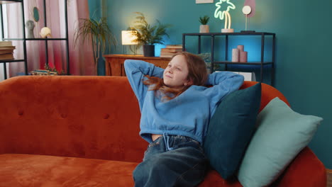 happy young child girl sitting comfortably with hands behind head relaxing on sofa in living room