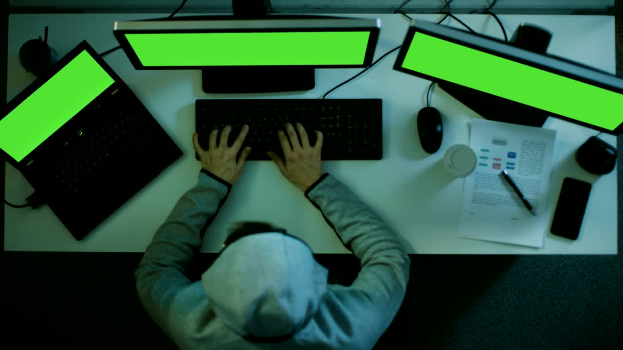 vista superior de un hacker entrando en un centro de datos internacional. sus dos monitores y portátil tienen pantalla verde. usa software malicioso hecho por él mismo.