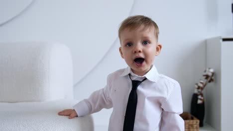 adorable caucasian toddler boy wearing white shirt and black tie stands indoors. cute baby climbs on the sofa.
