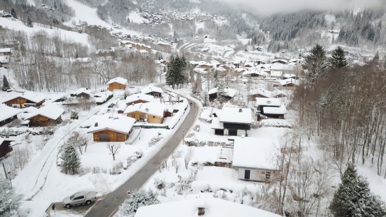 雪に覆われたスキー村を飛行するドローン撮影、低い雲、山の木々、シャレーを背景にカメラがパンアップする様子