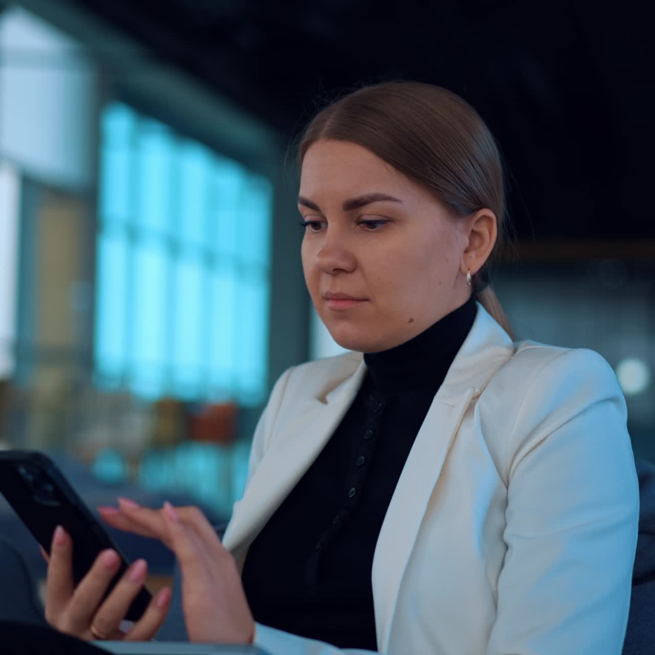 Business lady wearing white jacket sits in comfortable armchair focused on her phone. Caucasian woman working using her smartphone. Close up. Blurred backdrop
