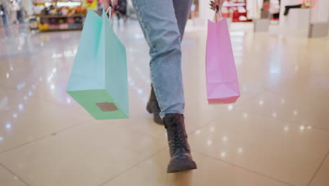 close-up leg view of person walking through a brightly lit shopping mall carrying pastel-colored shopping bags, stylish boots step confidently on polished floor reflecting retail lights