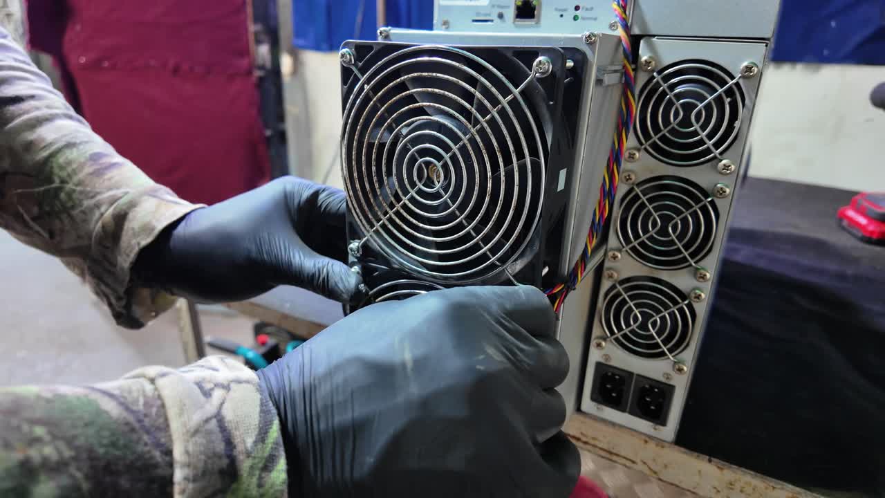 Close-up technician wearing black gloves while servicing or replacing cooling fan on a cryptocurrency mining machine. Multiple exhaust fans, power connectors, and wiring. Maintenance workspace