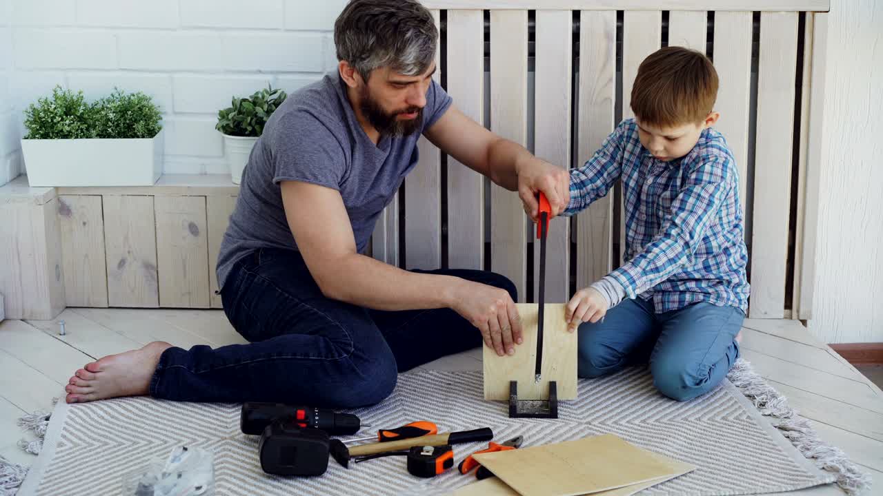el niño concentrado está aserrando una hoja de madera contrachapada con una sierra de mano y su padre está sosteniendo madera con sus manos y ayudando a su hijo a enseñarle. compartir concepto de experiencia.