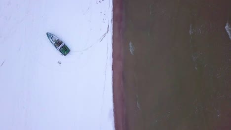 aerial birdseye view of baltic sea coast on a overcast winter day with green coastal fisherman boat, beach with white sand covered by snow, coastal erosion, wide angle drone shot moving forward