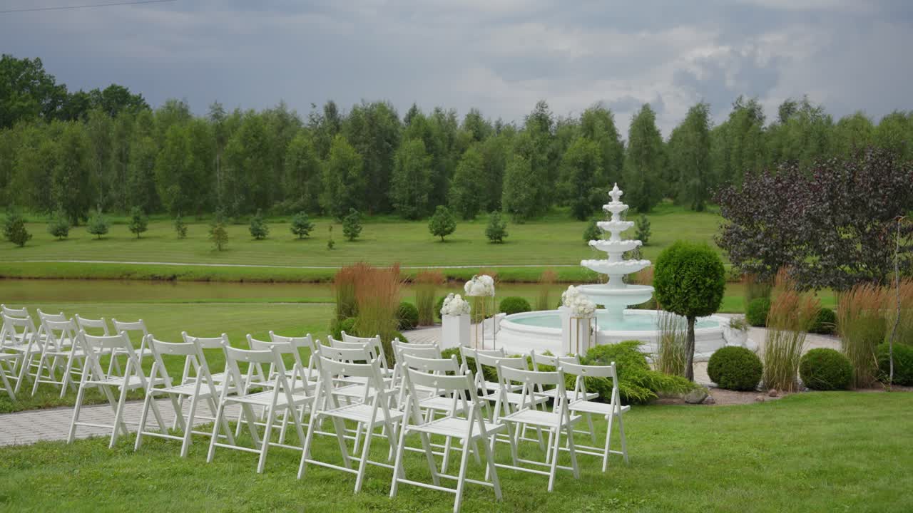 Outdoor wedding ceremony setup with white chairs and decorative fountain in a beautifully landscaped garden surrounded by trees and greenery under an overcast sky