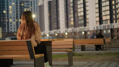 back shot of a girl with long brown hair in a peach jacket and black trouser, seated on a park bench. she is raising her head slightly, with tall,blurred buildings and distant people in the background