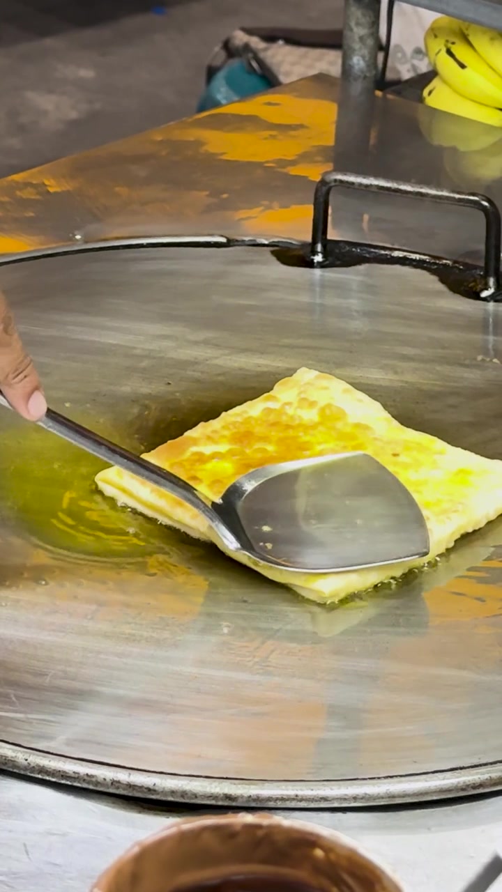 En gadesælger tilbereder dygtigt thailandsk roti på en varm stegeplade, hvilket fremviser traditionelle madlavningsteknikker i livlige markedsomgivelser.
