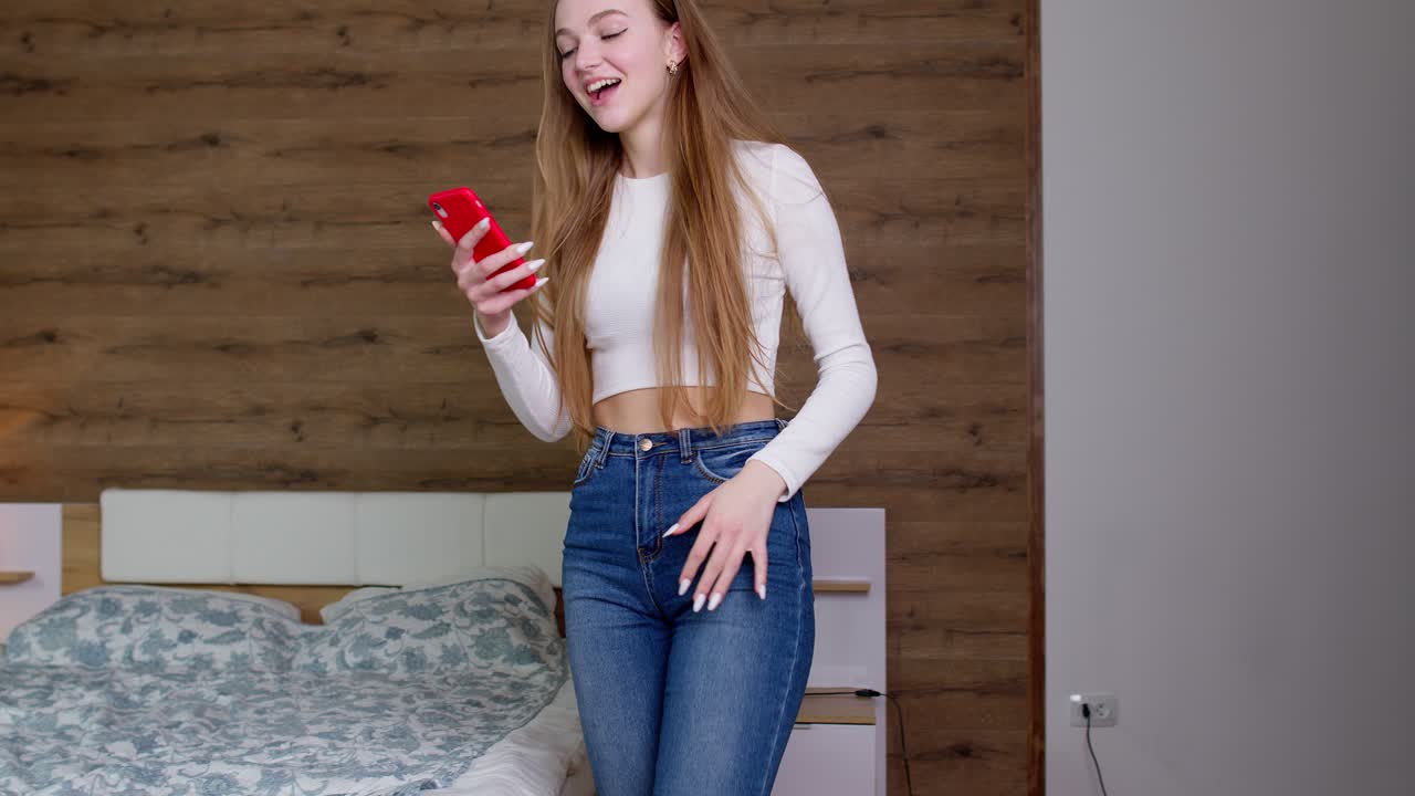 Giovane donna vincitrice, felicissima e sorridente, che salta e balla a casa festeggiando il successo di una vincita al telefono cellulare.