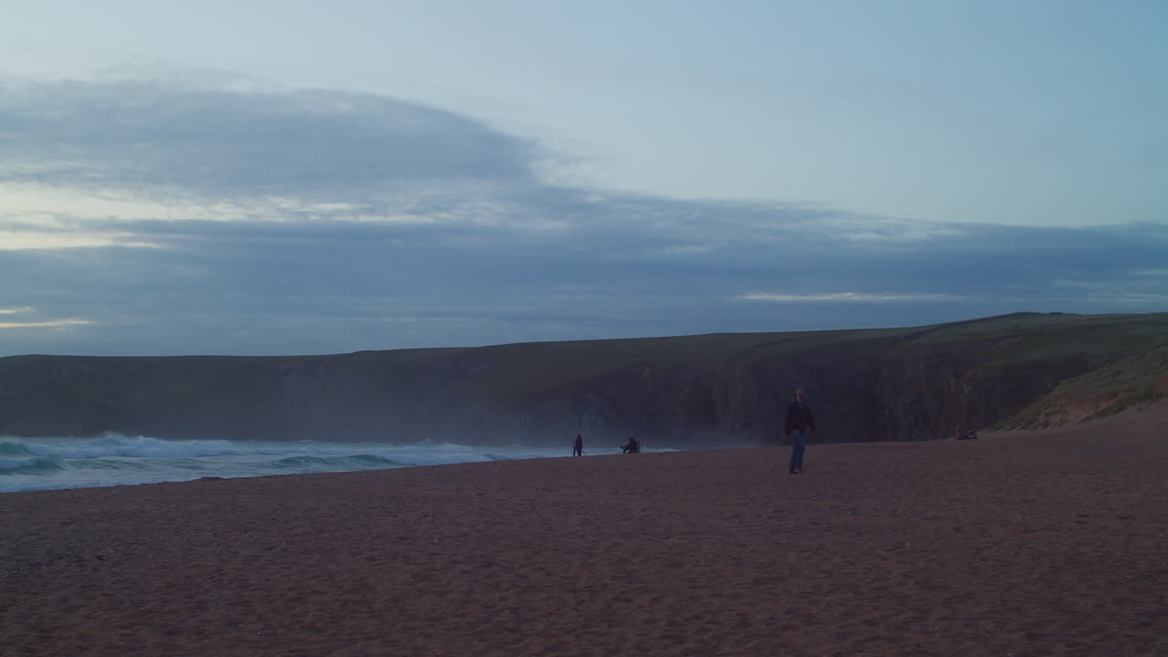 человек идет по песчаному пляжу с разбивающимися волнами в сумерках бухта холивелл, корнуолл, англия