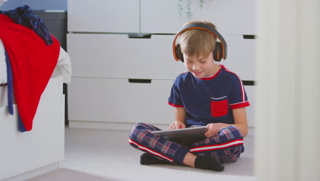 boy at home in bedroom wearing wireless headphones playing on digital tablet sitting on floor