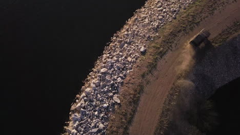 Quick drone flyover view of an old pickup truck driving around a curve on a jetty sea wall breakwater at sunset
