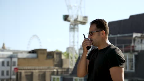 hombre con una camiseta negra usando walkie-talkie en un techo con una vista clara de un horizonte urbano con edificios y vegetación, mostrando la comunicación en un entorno urbano