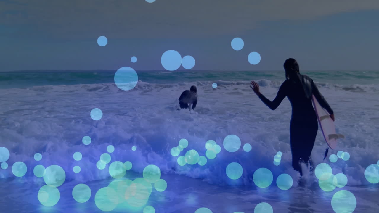 Two surfers paddling and wading into foamy ocean waves, showcasing tech using blue circle overlays