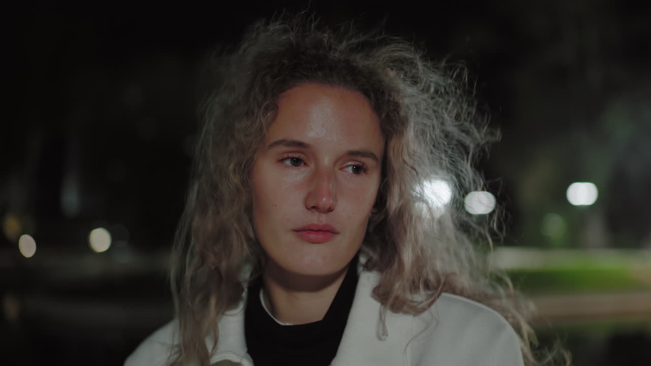 Closeup of calm girl standing alone at night with gentle wind blowing through her hair, eyes shifting slowly open in quiet reflection, illuminated by blurred glowing background streetlights
