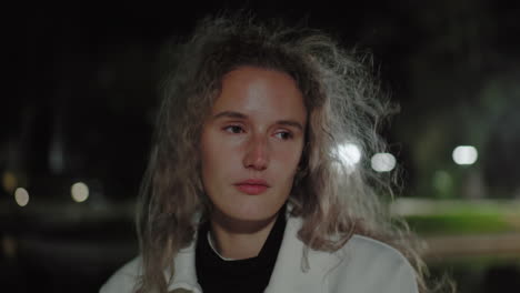 closeup of calm girl standing alone at night with gentle wind blowing through her hair, eyes shifting slowly open in quiet reflection, illuminated by blurred glowing background streetlights