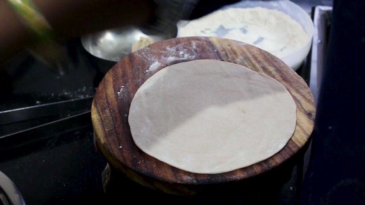 Close-up tangan menggulung adonan chapati di atas papan chakla yang ditaburi tepung. Ideal untuk dokumenter makanan, tutorial memasak, masakan India, dan proyek gaya hidup budaya.