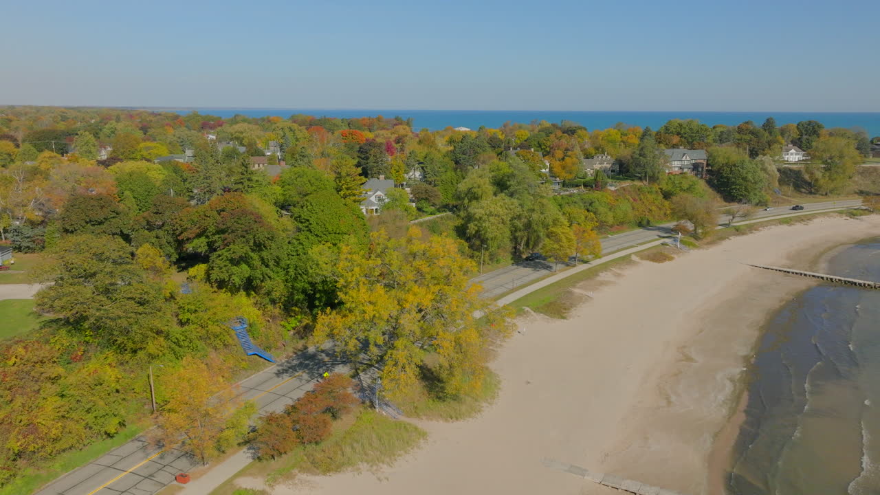 Vista aérea con dron sobre la playa del lago Michigan en dirección a una carretera costera con coches, casas en un barrio y coloridos árboles otoñales bajo un cielo azul despejado en un hermoso día de otoño