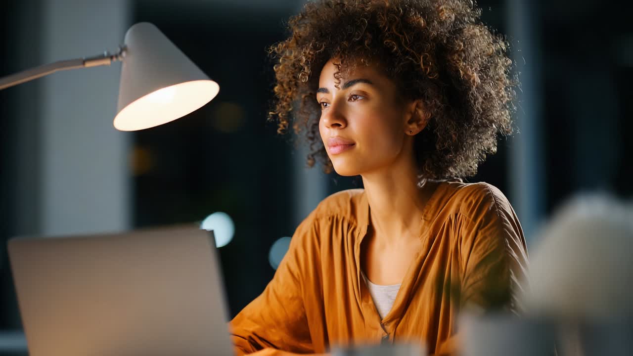A Focused Young Woman with Curly Hair Working Late at Night at Her Desk, Illuminated by a Desk Lamp, Absorbed in Front of a Laptop, Symbolizing Dedication and Productivity in a Cozy Home Office Setting