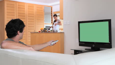 man watching the television while his wife is preparing a meal