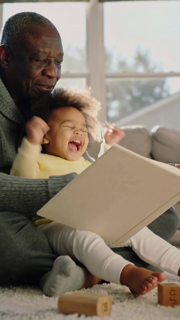 Video verticale: Uomo che appoggia un libro e aiuta un bambino a girare le pagine su un tappeto a casa, vicino a blocchi e una maglia grigia