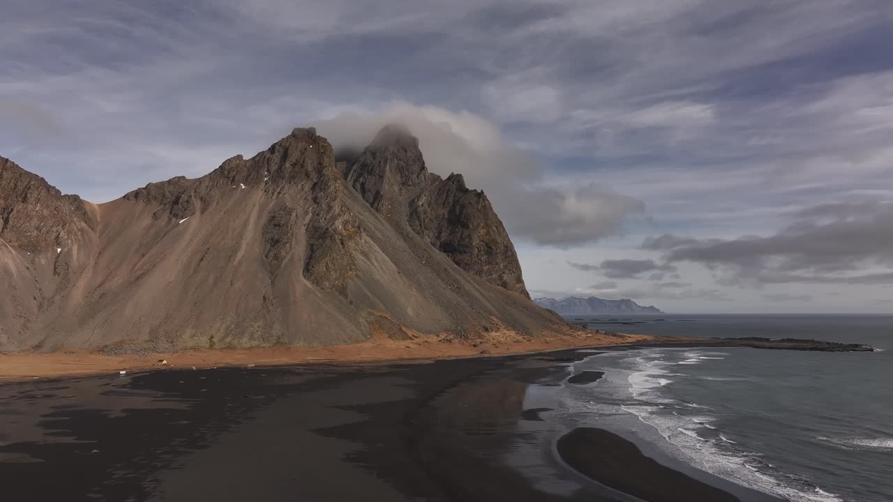 Wide drone view of Vestrahorn’s jagged peaks meeting the ocean, with black sand beaches and dramatic skies at Stokksnes, Iceland.