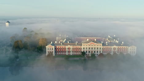 Toma-Aérea-Revelada-Del-Majestuoso-Edificio-Del-Castillo-Del-Palacio-De-Jelgava-Entre-Un-Denso-Día-De-Niebla-En-Letonia