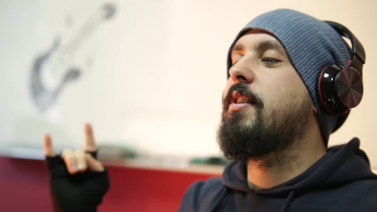 Young hipster male with a beard and hat under wireless headphones is looking at the camera and gesturing sign of rocker