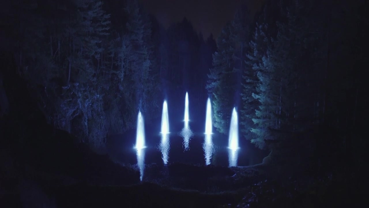 Wide shot of a Blue lit Fountain on lake surrounded by forest, Tilt up, Night