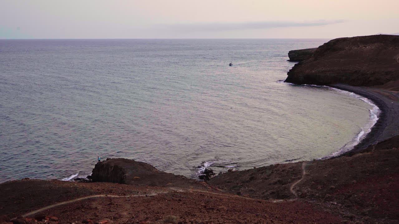 una toma estática de una playa salvaje en las islas canarias de españa con un bote flotando en el horizonte y un hombre pescando en la esquina inferior izquierda